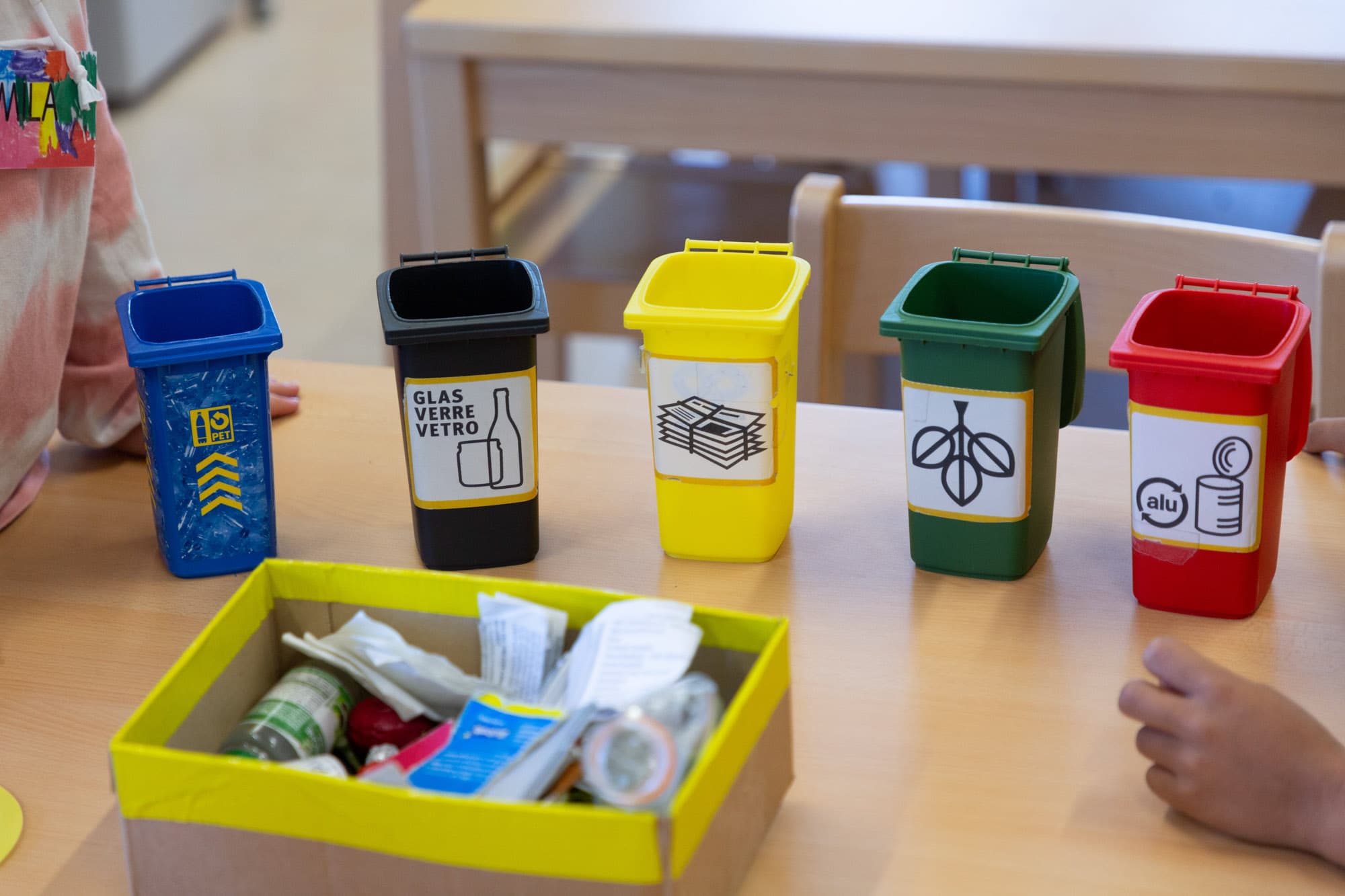 Fünf verschiedenfarbige Mini-Plastikkübel stehen symbolisch für verschiedene Recyclingtonnen auf einem Tisch.