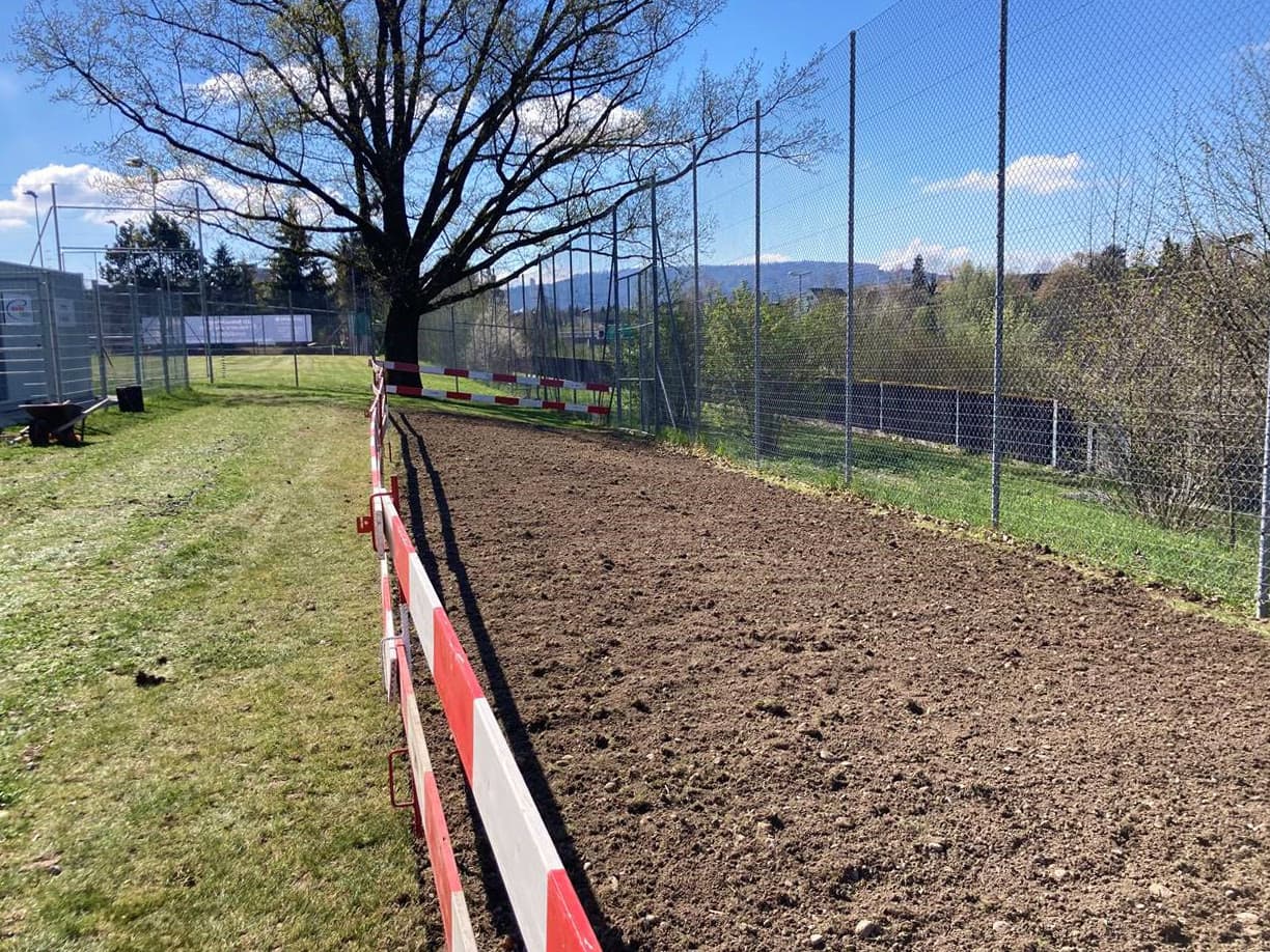 Mit Bauabsperrungen eingezäunte Fläche bei einem Sportplatz, auf der früher Rasen stand und die umgegraben und für eine Blumenwiese vorbereitet wurde