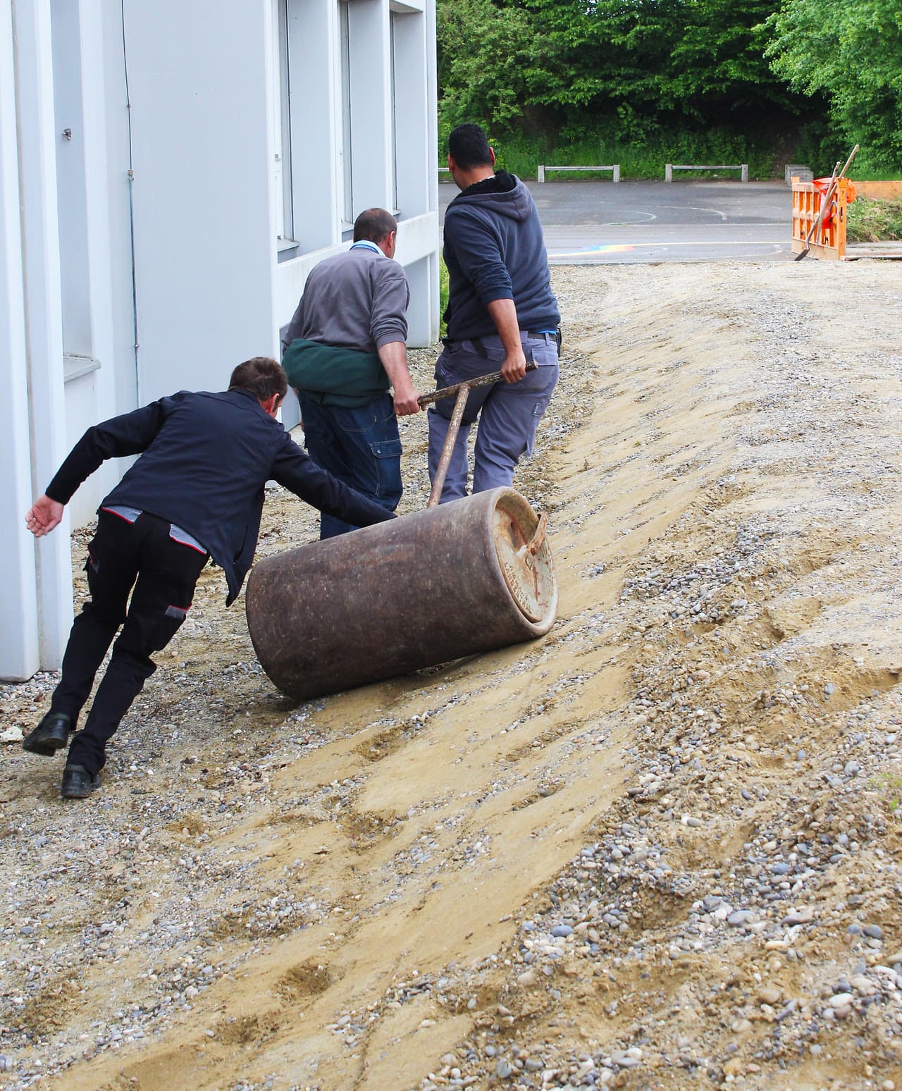 Drei Männer stossen eine Walze über eine Fläche mit Sand, Erde und Kies