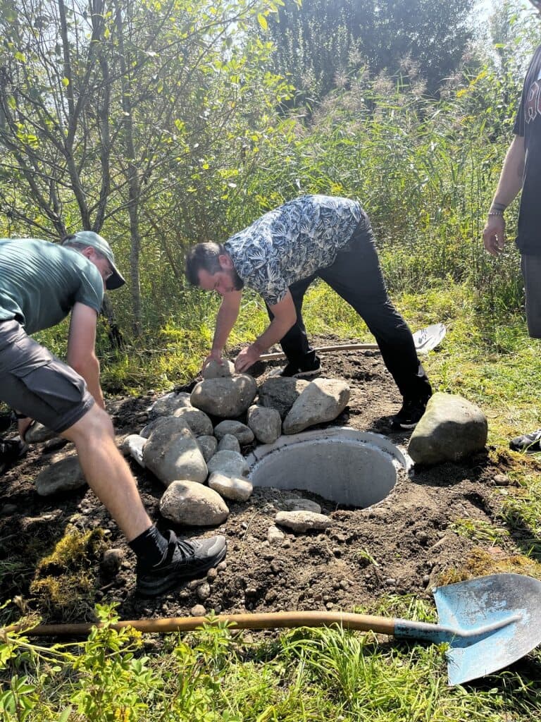 Zwei Männer stapeln Steine um ein eingegrabenes Betonrohr, um eine Unkenwanne zu erstellen.