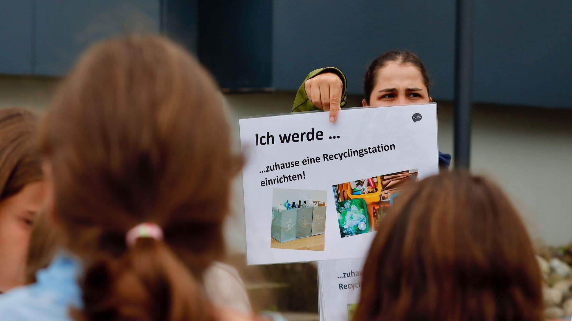 Eine Lehrperson hält ein Plakat hoch, auf dem steht «Ich werde zuhause eine Recyclingstation einrichten!». Vor ihr stehen mehrere Schüler:innen und hören zu. Auf dem Plakat sind Bilder von Recycling-Behältern abgebildet.