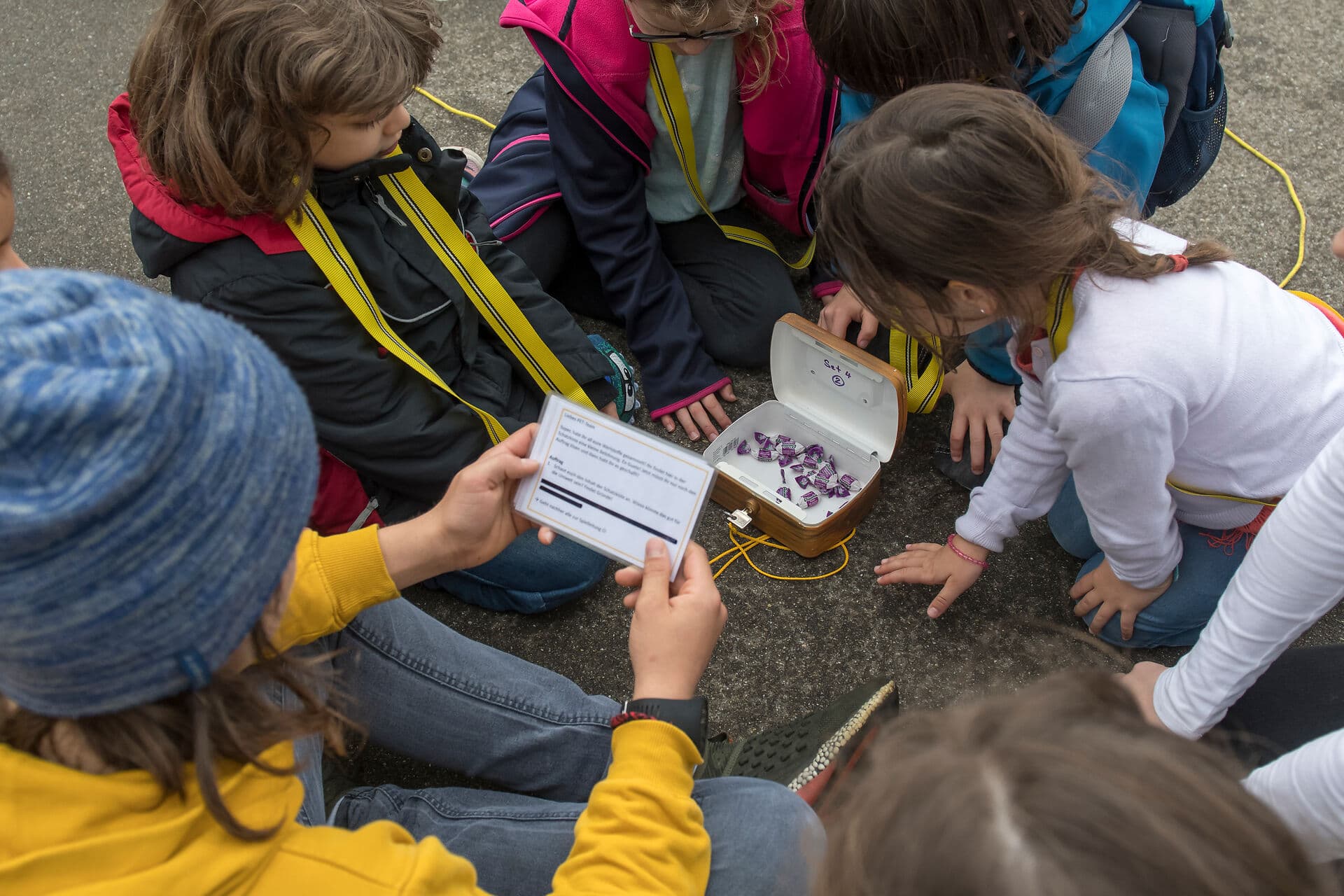 Kinder sitzen auf dem Pausenplatz rund um eine Schatulle gefüllt mit Ricola-Bonbons und lesen eine Spielinstruktion